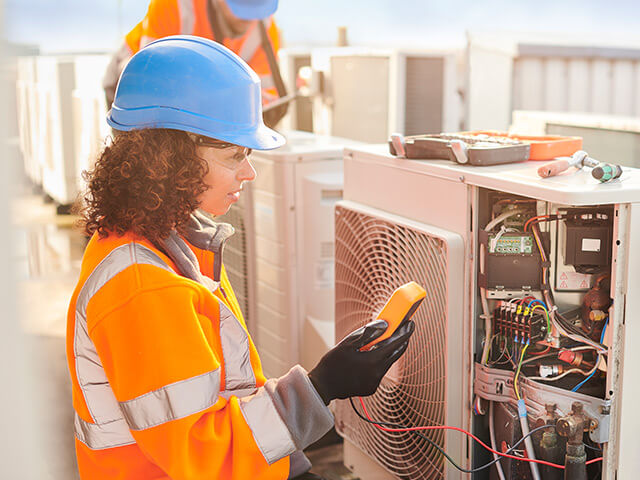 Woman with blue hardhat and orange vest working on HVAC unit using diagnostic materials