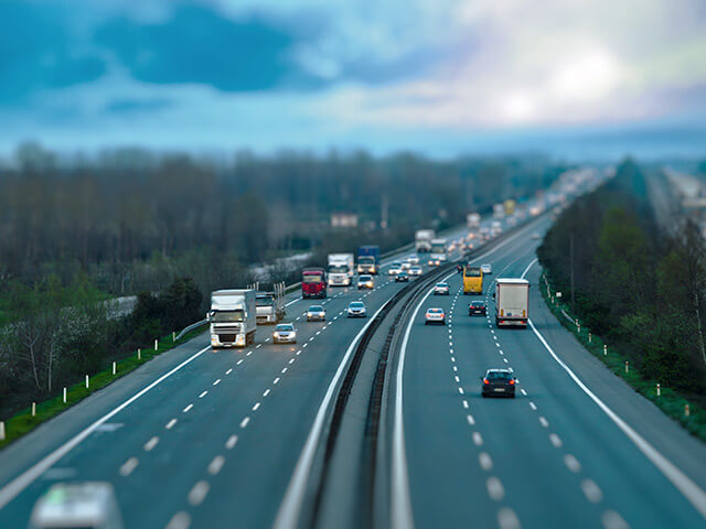 Cars traveling on six lane highway with blue and purple colored sky, green grass, and trees